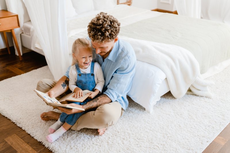 Man smiling while reading storybook to girl on lap as she laughs