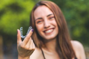 Smiling woman with brown hair and purple nails holding clear aligner tray in the foreground in front of a blurry green background