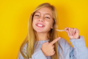 Girl in grey sweater holding clear aligner in front of a yellow background